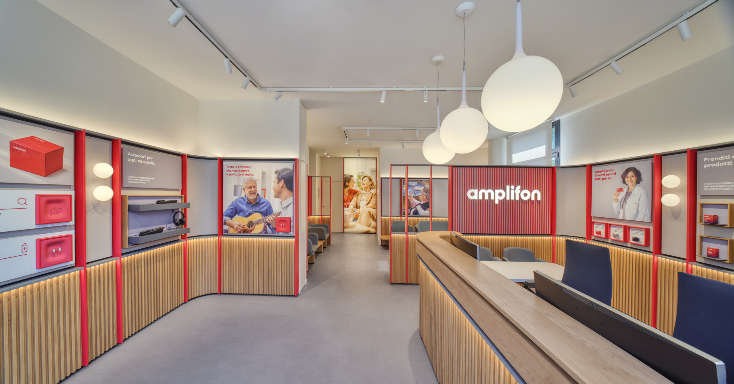 Interior of the full-scale replica Amplifon store at headquarters: waiting area with seating and a backlit brand wall, a wooden counter, and a product display wall.
